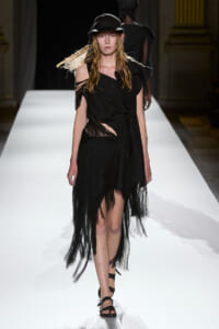 Model walks the runway in a black fringed dress with shredded hem and a black cap, spotlighted on a white catwalk.