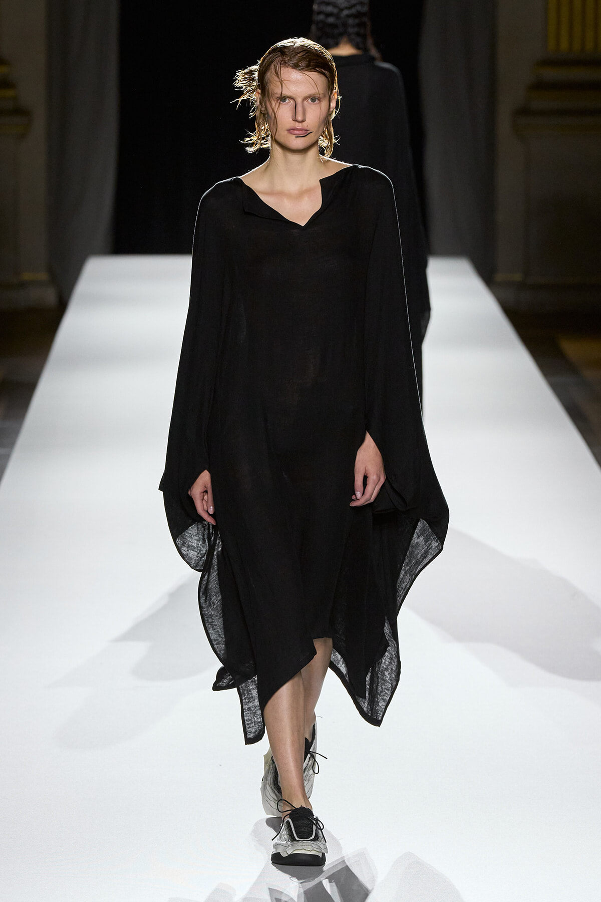 Model in a loose black sheer dress walks down a white fashion runway on a catwalk with an attentive expression.