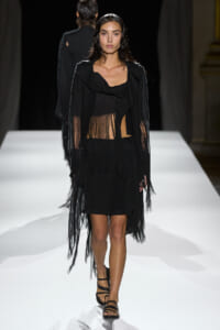 Model walking the runway in a sheer black fringe dress with a cape-like outer layer and strappy sandals.