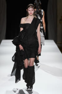 Model walks the runway in a black, draped, one-shoulder dress with a long, wrapped train and matching black boots.