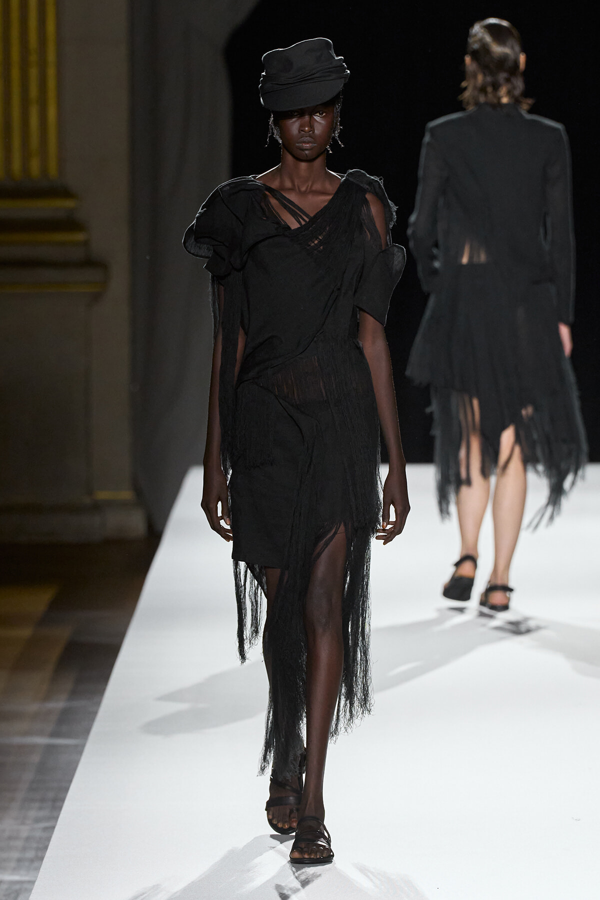 Model walking a fashion runway in an all-black, fringed dress with a matching black hat and flat sandals on a white catwalk.