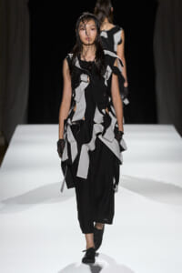 Model walks the runway in a black dress with layered gray fabric strips and wet hair.