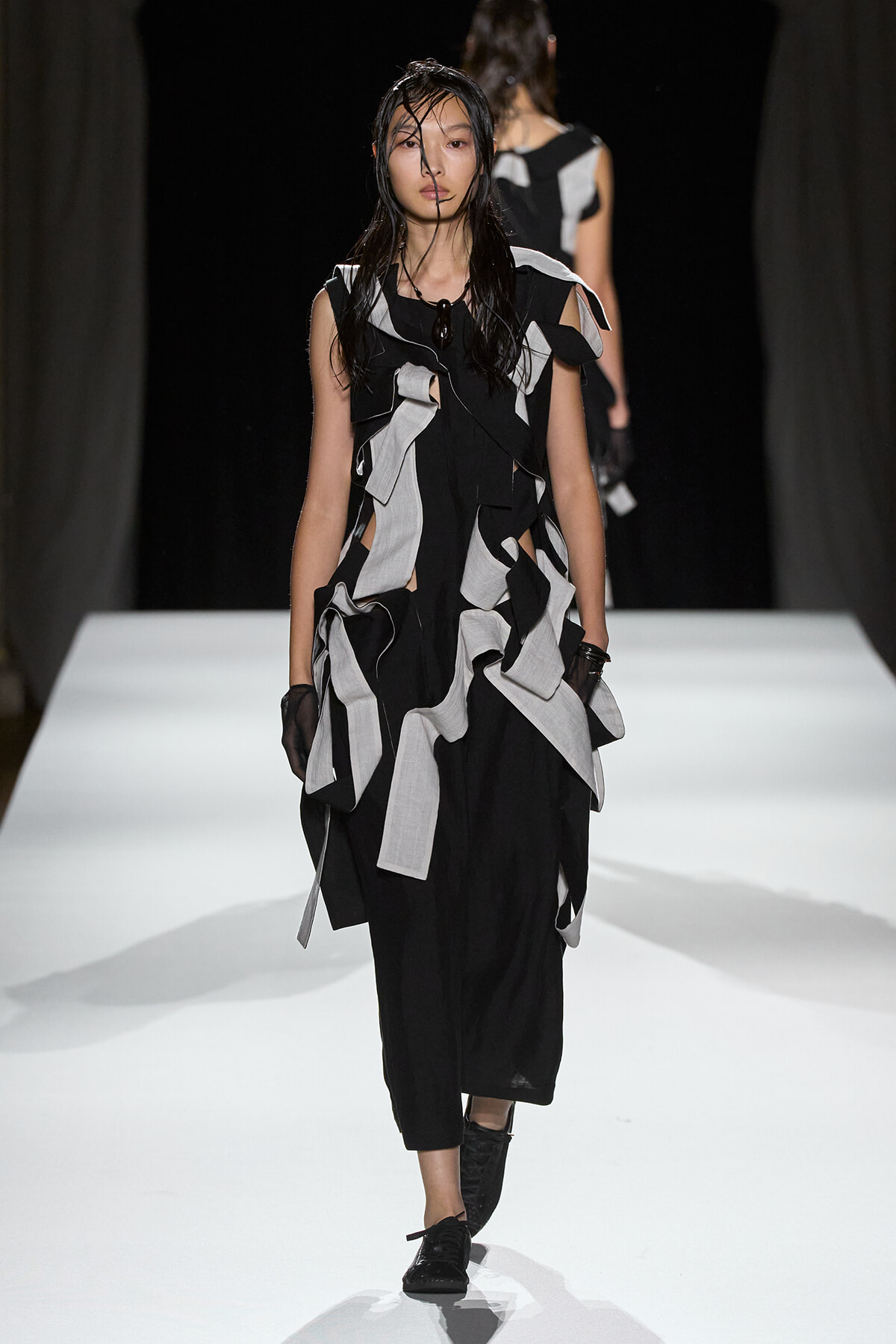 Model walks the runway in a black dress with layered gray fabric strips and wet hair.