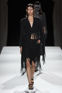 Model walking a runway in a black fringed blazer and skirt with strappy sandals.