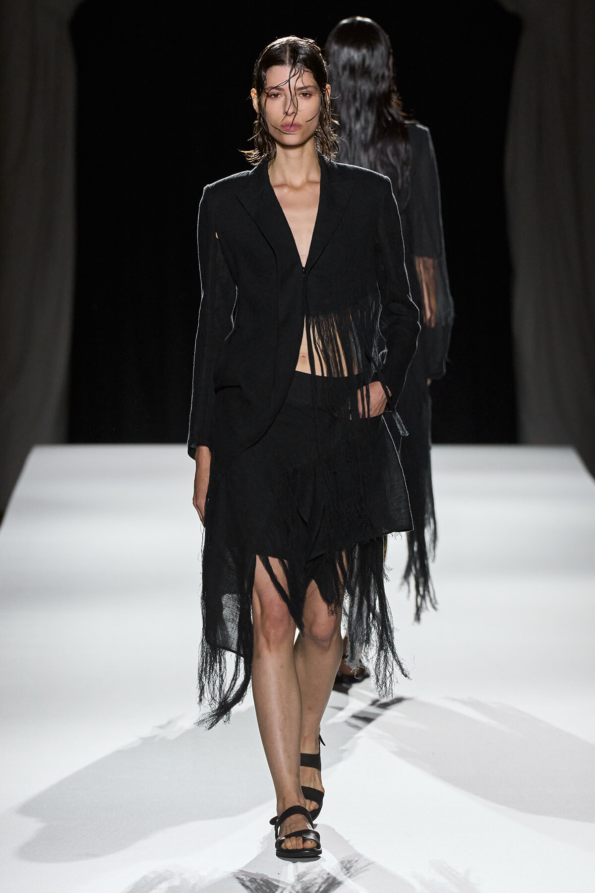 Model walking a runway in a black fringed blazer and skirt with strappy sandals.