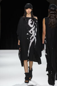Female model in an oversized black caftan with a white abstract print walks the runway, wearing a black cap and chunky black sandals; another model appears in the background.