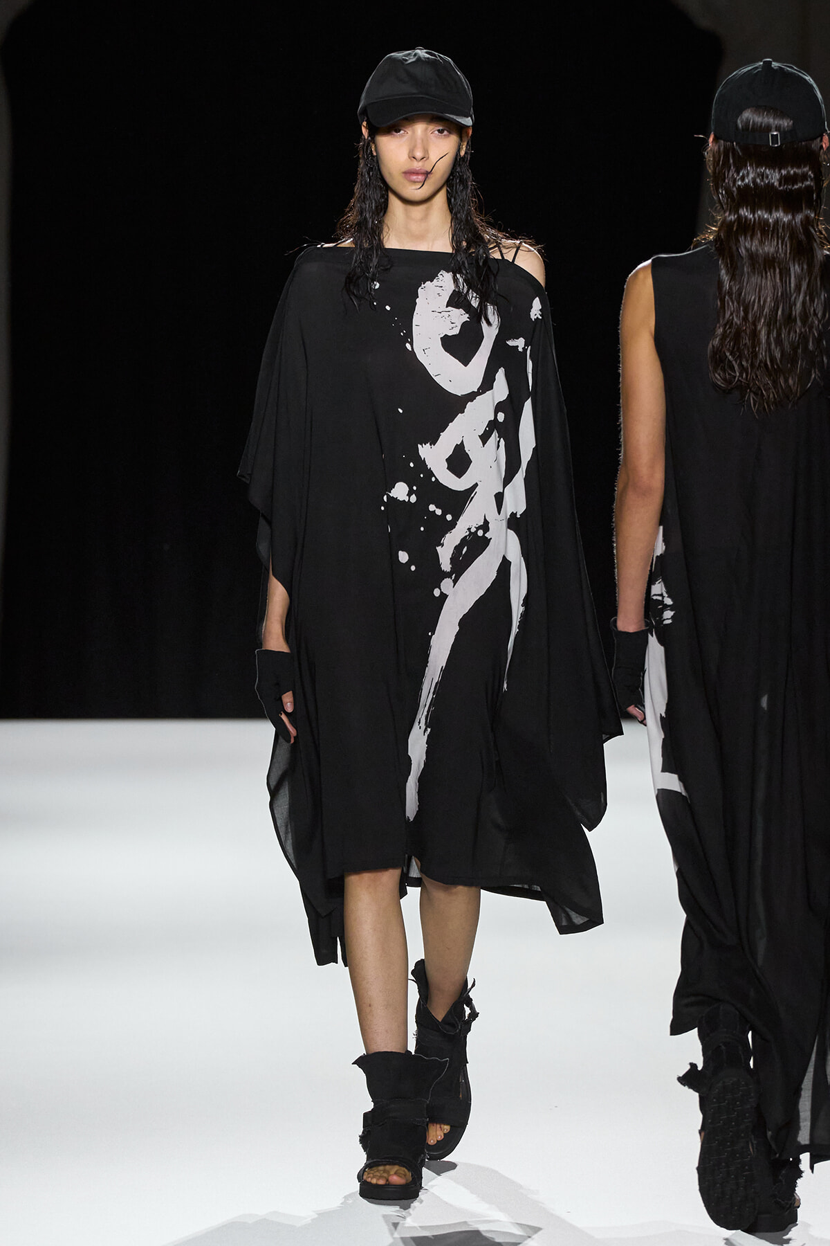 Female model in an oversized black caftan with a white abstract print walks the runway, wearing a black cap and chunky black sandals; another model appears in the background.