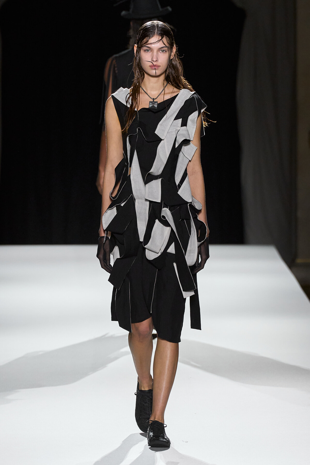 Model walks the runway in a black-and-white geometric patchwork dress made of ragged fabric strips.