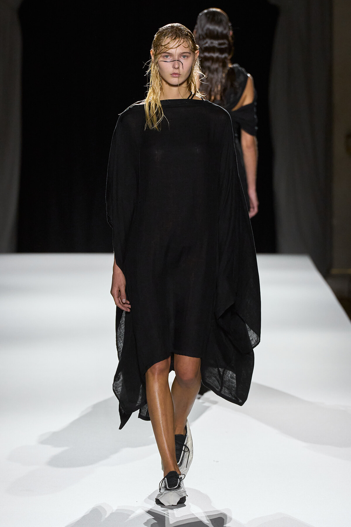 Model walking the runway in a loose black, poncho-style dress with an asymmetrical hem and white sneakers; wet, slicked-back hair and a focused expression.