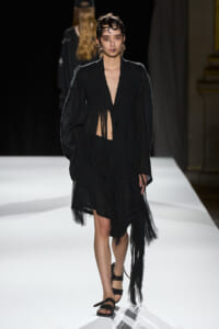 Female model on a white runway wearing a black oversized fringed blazer, matching black skirt, and strappy sandals.