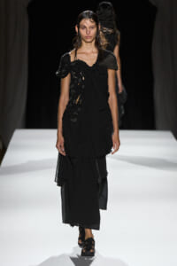 Model walking the runway in a black layered dress with irregular cutouts and a ruched bodice, with wet slicked-back hair.