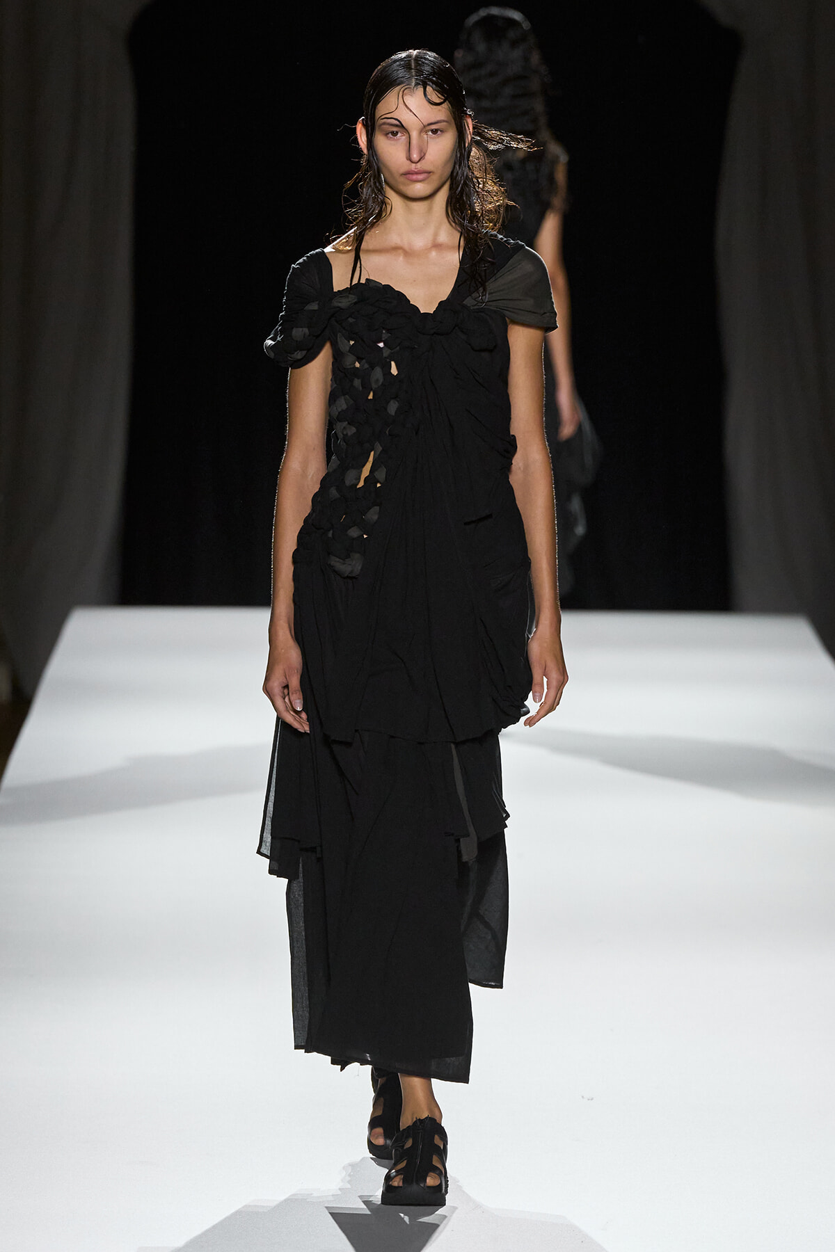 Model walking the runway in a black layered dress with irregular cutouts and a ruched bodice, with wet slicked-back hair.