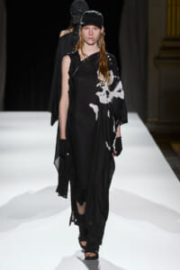 Model walks a black, draped dress with white abstract print on a white runway, wearing a black cap and gloves.