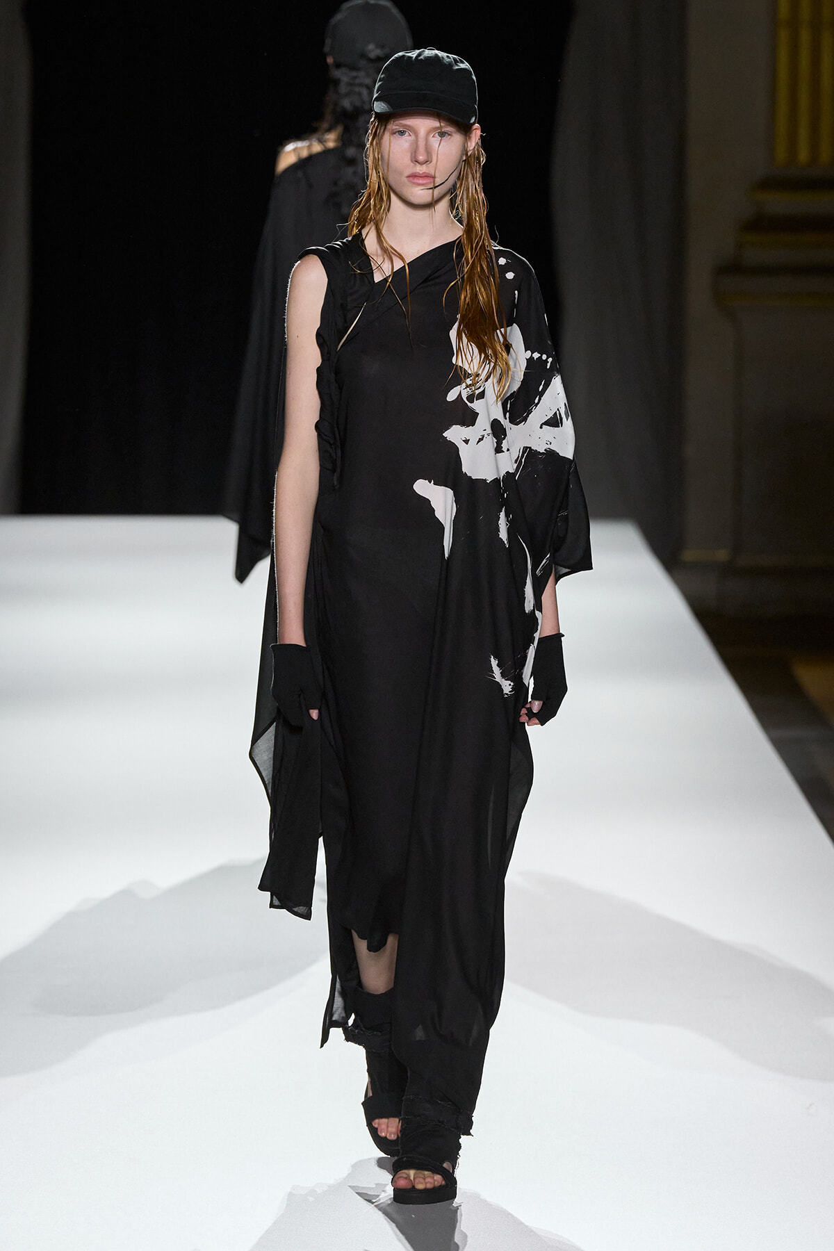 Model walks a black, draped dress with white abstract print on a white runway, wearing a black cap and gloves.