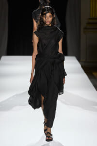 Model walks the runway in a black draped dress with a knotted waist, wearing black sandals and wet-looking hair on a white catwalk.