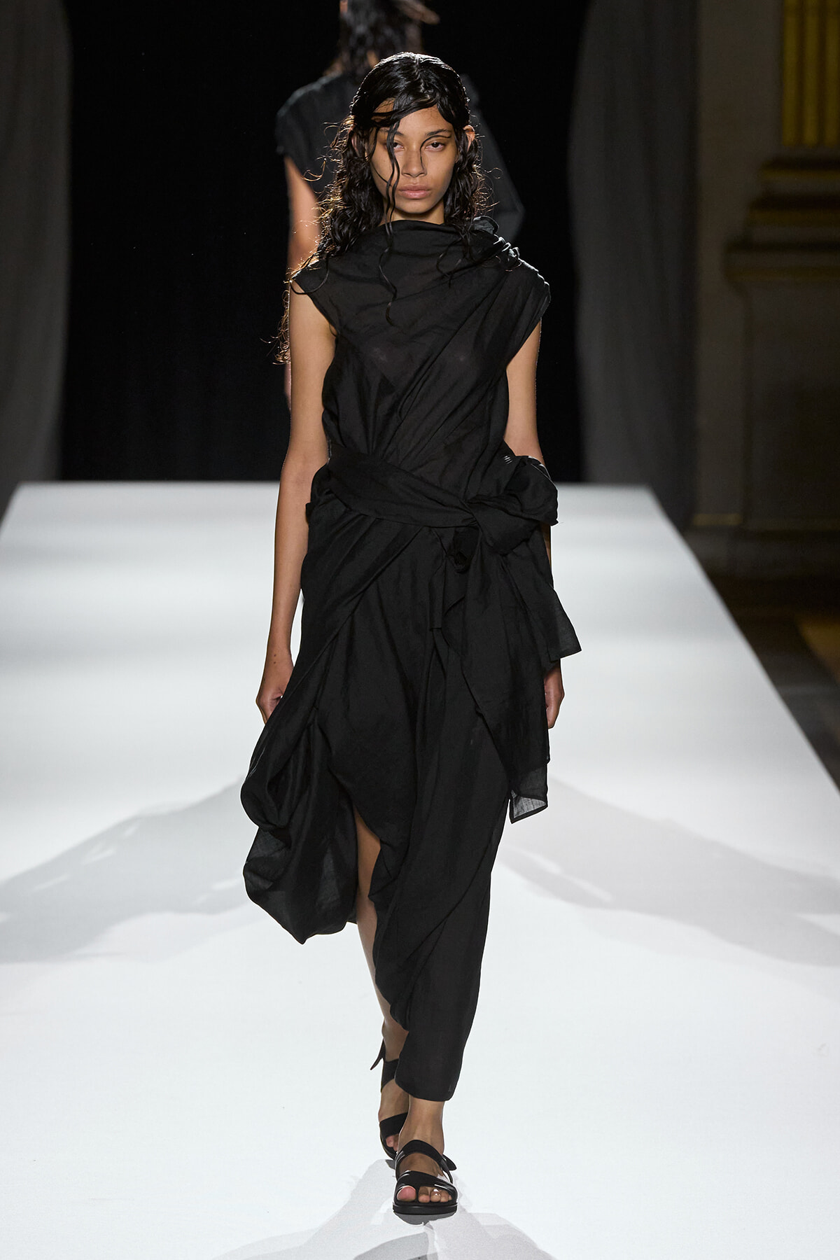 Model walks the runway in a black draped dress with a knotted waist, wearing black sandals and wet-looking hair on a white catwalk.