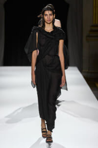 Model walking a white runway in a black draped one-shoulder outfit with strappy sandals.
