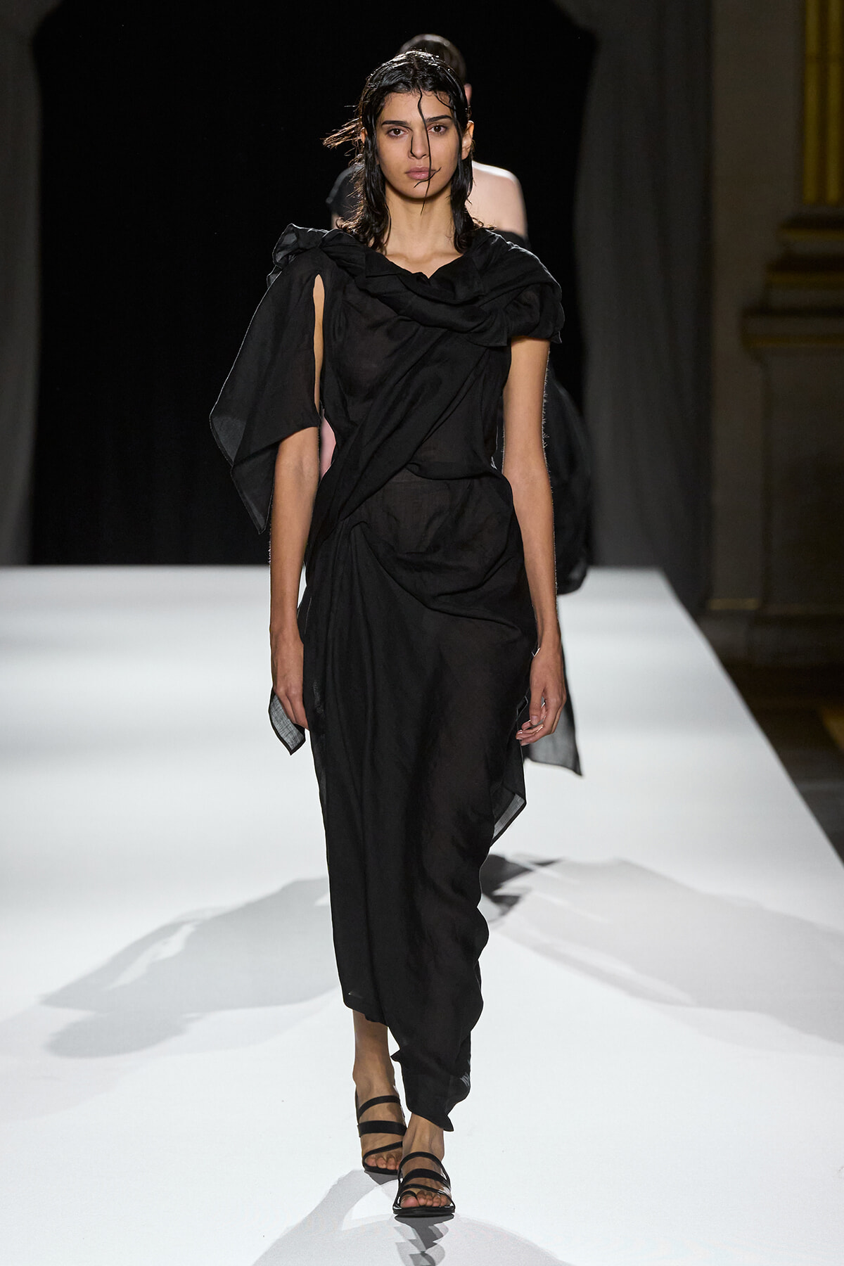 Model walking a white runway in a black draped one-shoulder outfit with strappy sandals.