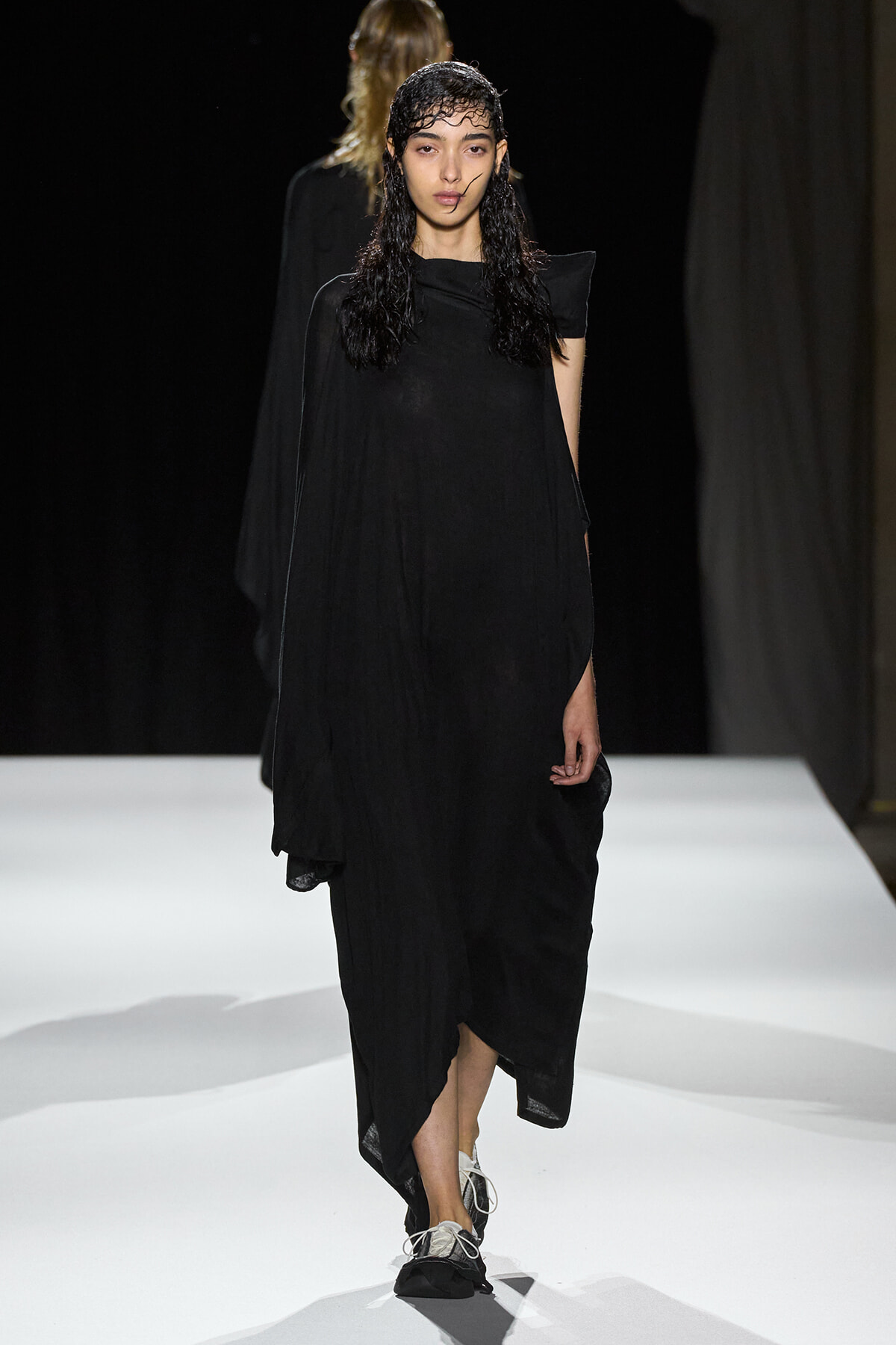 Model walks a dark, flowing black dress down a white runway, wet hair and minimalist makeup creating a moody look.