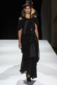 Model walking the runway in a layered black off-shoulder gown with ruffled panels and slicked-back hair on a white catwalk.