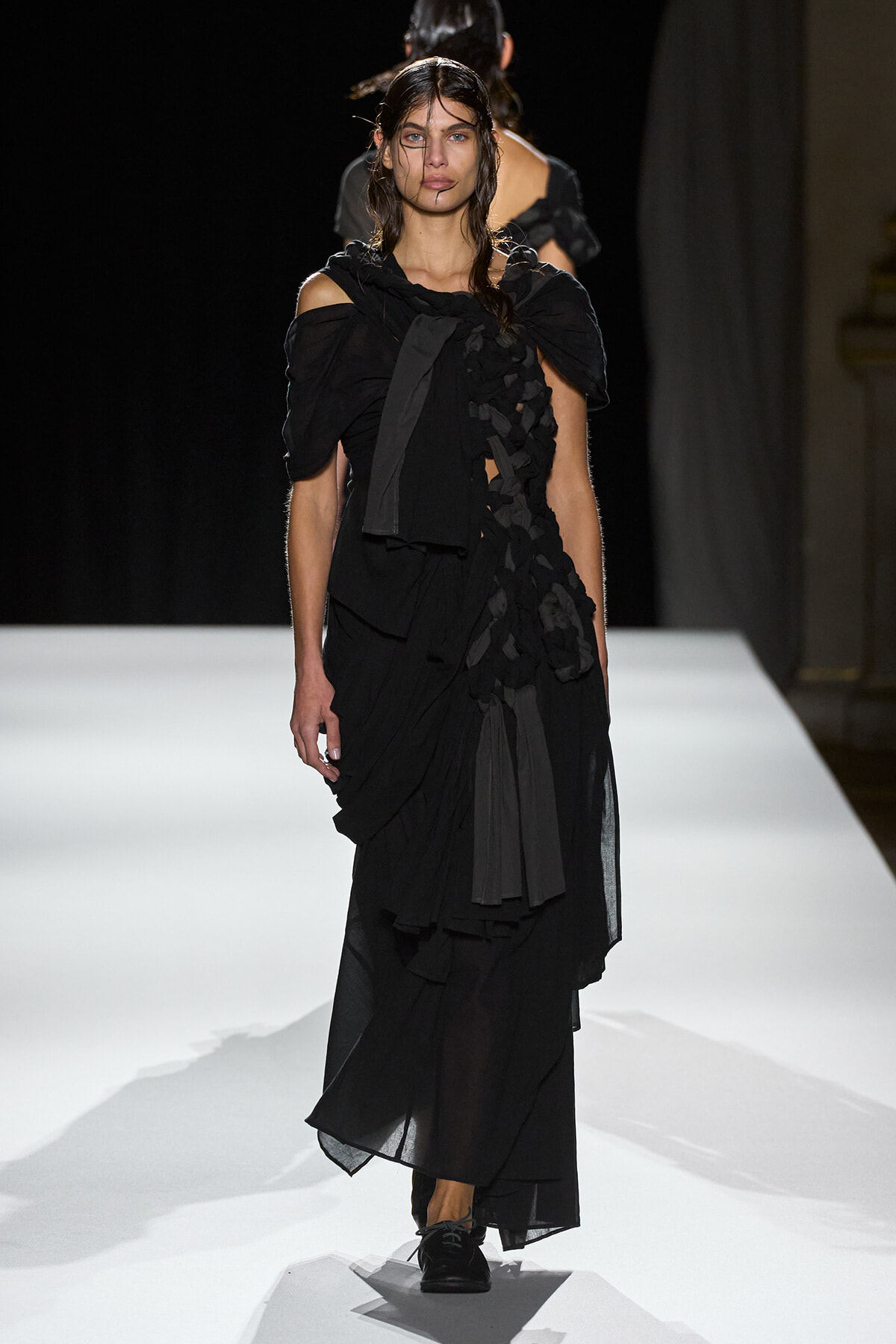 Model walking the runway in a layered black off-shoulder gown with ruffled panels and slicked-back hair on a white catwalk.