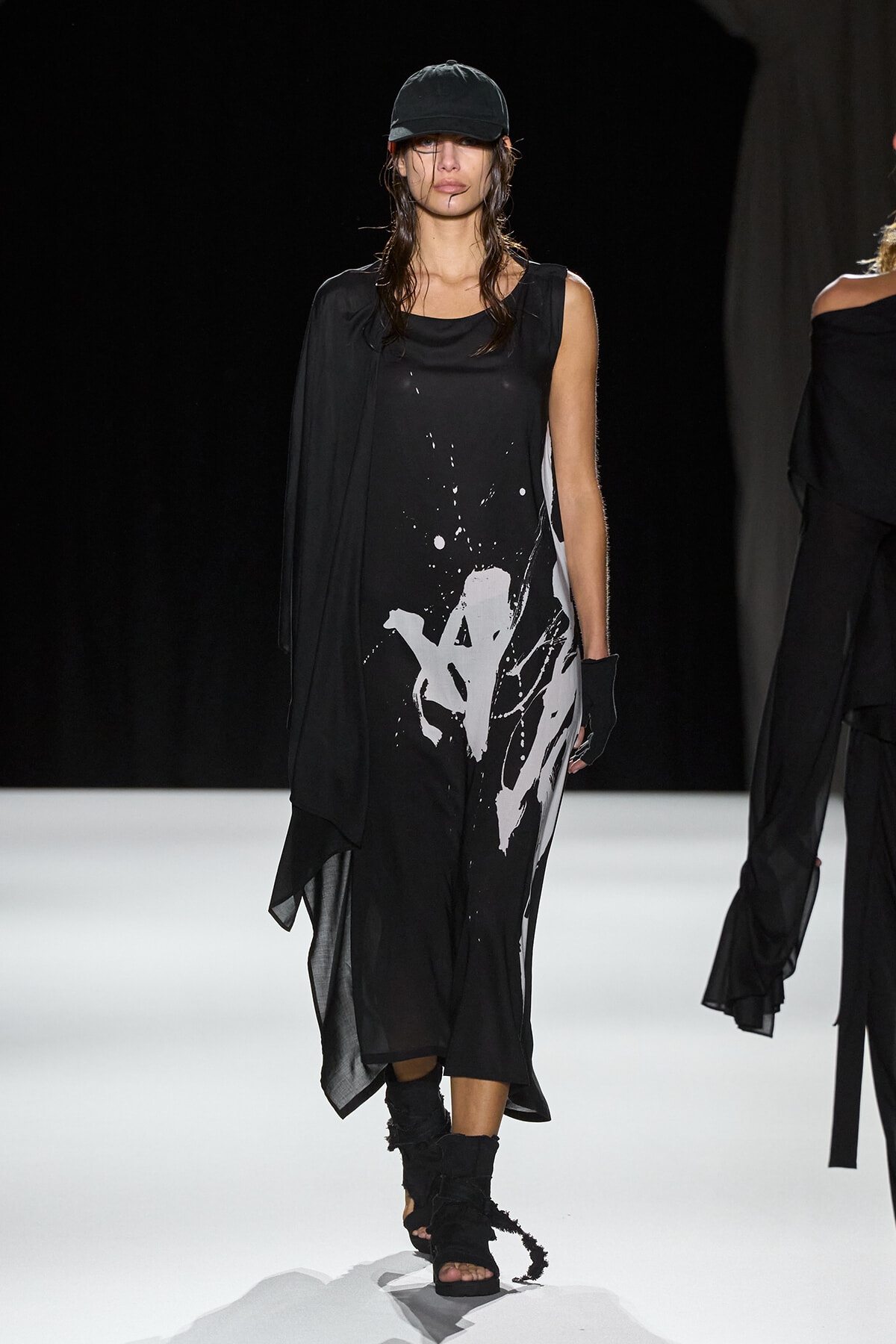 Model on a fashion runway wearing a black sleeveless dress with a white paint-splatter design, black cap, and jagged black sandals with gloves, walking forward.