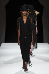 Female model walks the runway in a black sleeveless button-front dress with fringe strands and a wide-brimmed hat with feathered accents.