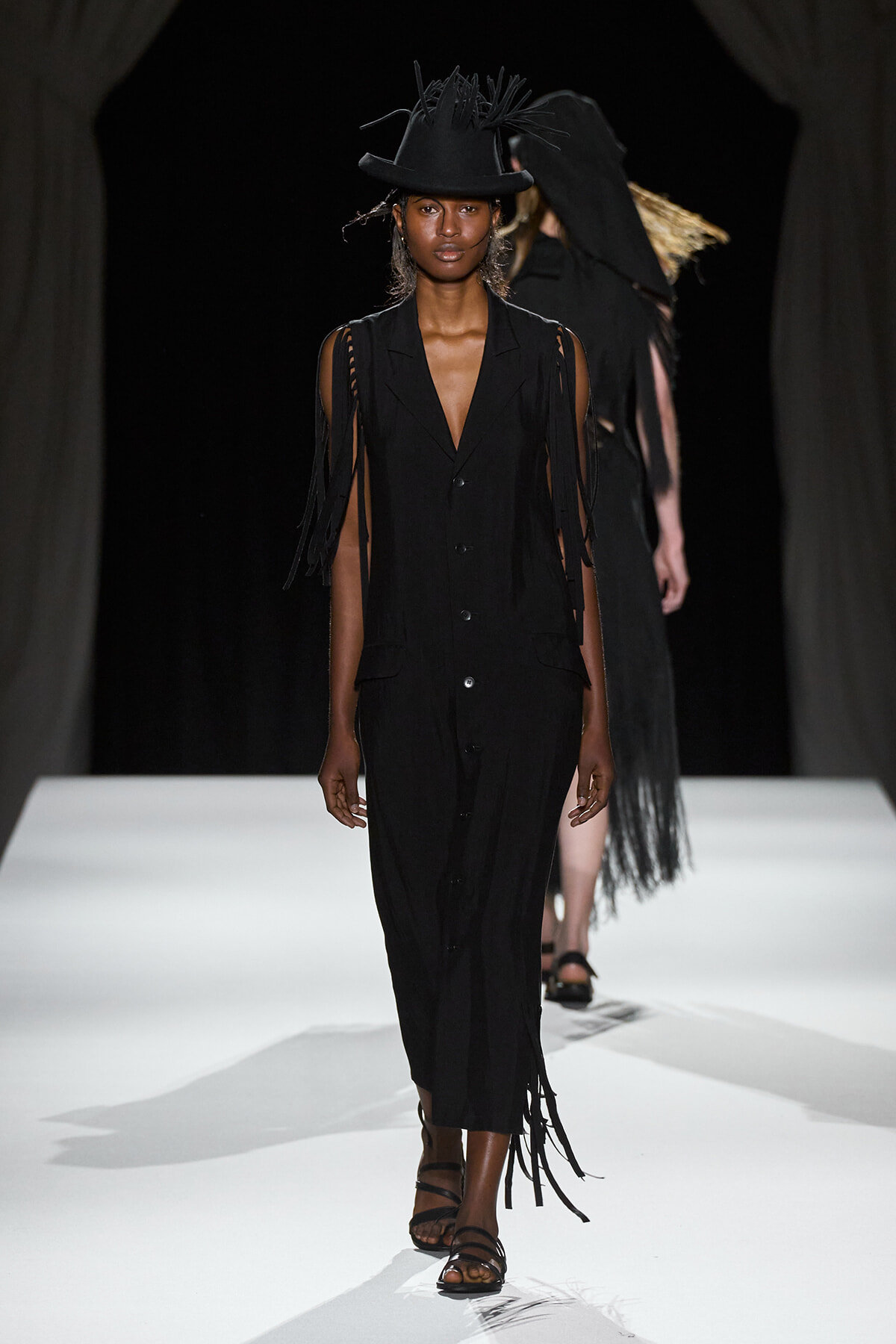 Female model walks the runway in a black sleeveless button-front dress with fringe strands and a wide-brimmed hat with feathered accents.