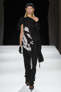 Model walking the runway in a black, one-shoulder dress with white abstract print and a cap, gloves, and open-toe sandals.