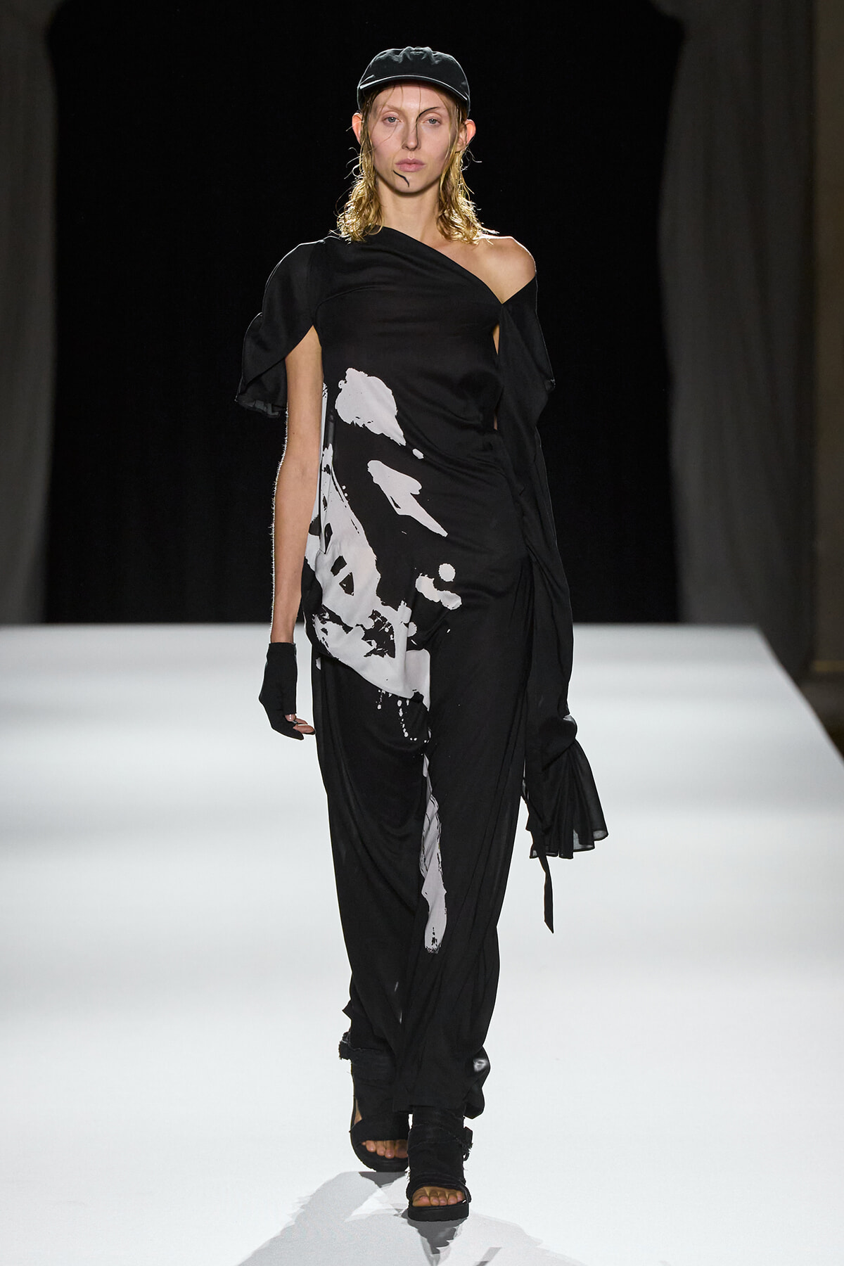 Model walking the runway in a black, one-shoulder dress with white abstract print and a cap, gloves, and open-toe sandals.