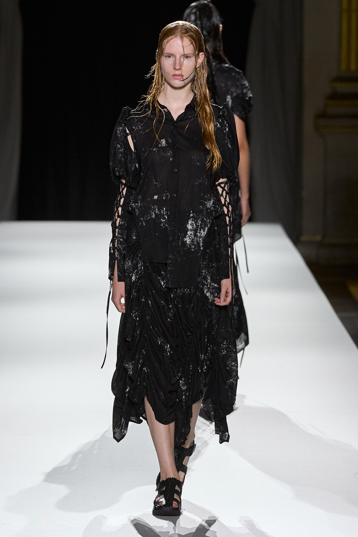 Female model walks a white runway in a black, splattered, oversized dress with lace-up sleeves and draped skirt.