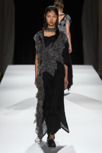 Female model walking a runway in a black dress with a layered gray lace overlay and sheer accents