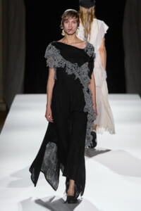 Model walking a runway in a black dress with gray lace appliqué cascading diagonally from shoulder to hem.