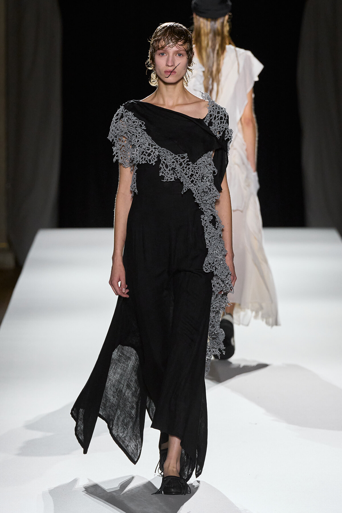 Model walking a runway in a black dress with gray lace appliqué cascading diagonally from shoulder to hem.