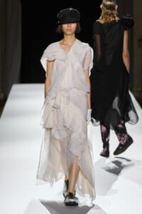 Female model walks a white layered chiffon gown down a runway, wearing a black cap and sneakers in a fashion show venue.