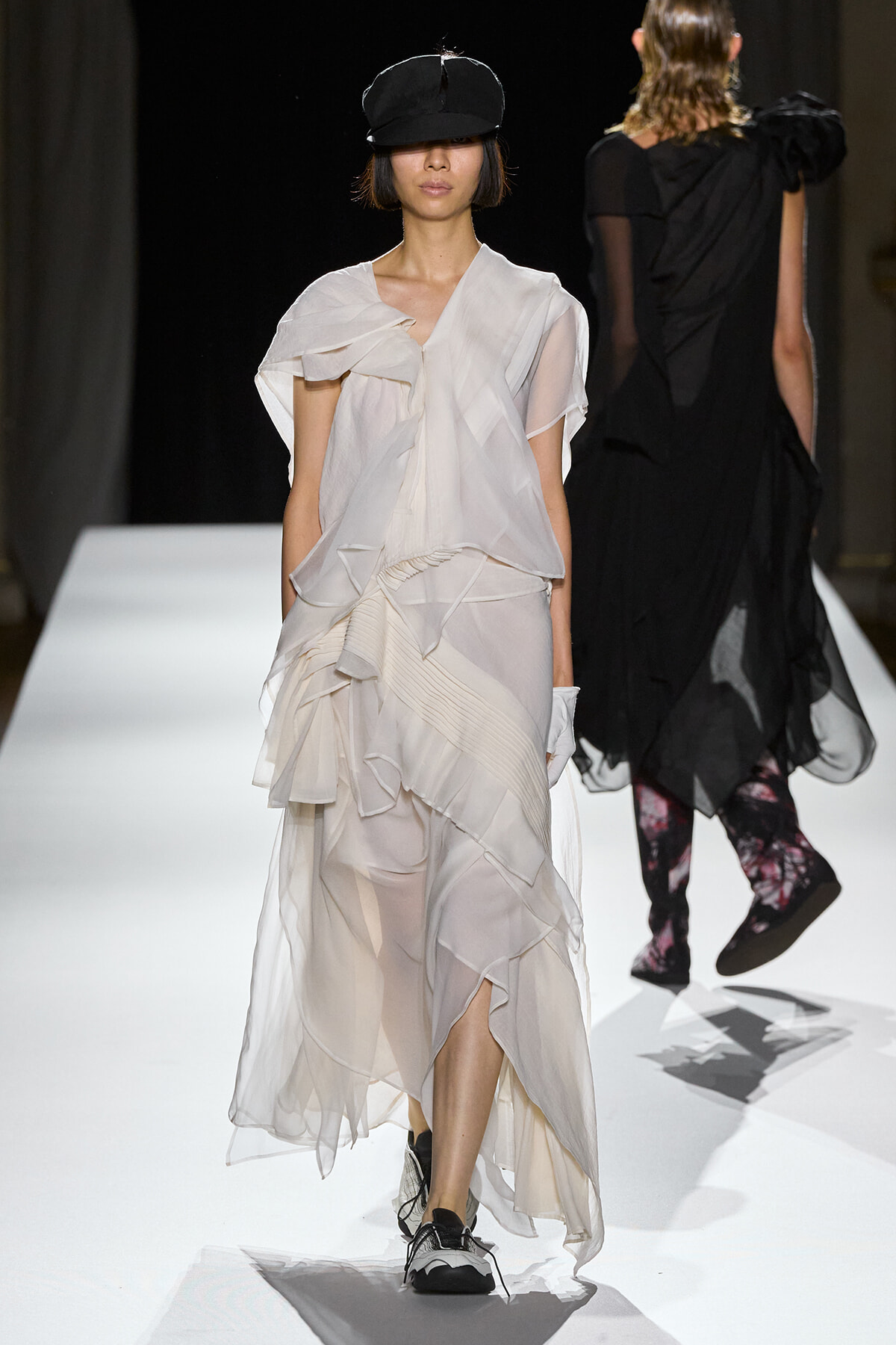Female model walks a white layered chiffon gown down a runway, wearing a black cap and sneakers in a fashion show venue.