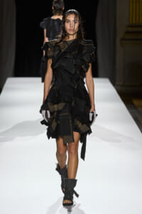 Model walks a white runway wearing a black, multi-layered ruffle dress with olive accents and matching gloves.