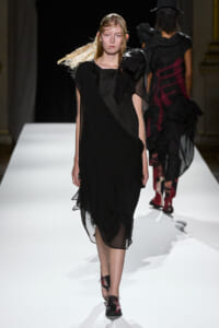 Female model walking a fashion runway in a black, layered, semi-sheer dress with a prominent shoulder bow/ruffle detail.