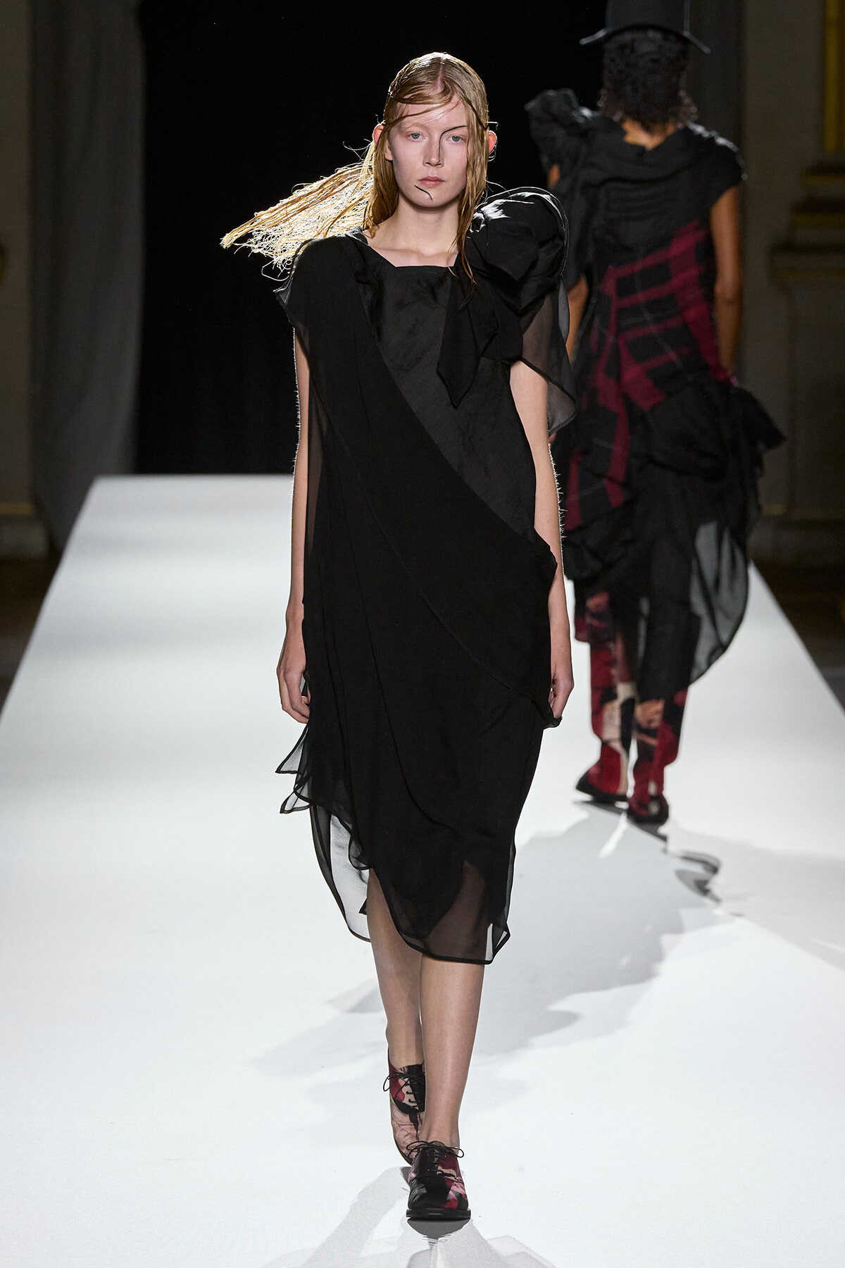 Female model walking a fashion runway in a black, layered, semi-sheer dress with a prominent shoulder bow/ruffle detail.