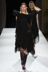 Female model walks a white runway in a black, layered dress with sheer panels and patterned knee-high boots.