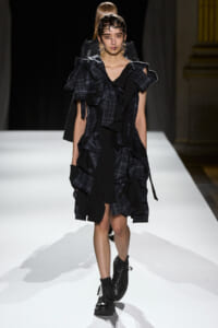 Model walks the runway in a dark plaid, ruffled dress with oversized shoulders and black combat boots.