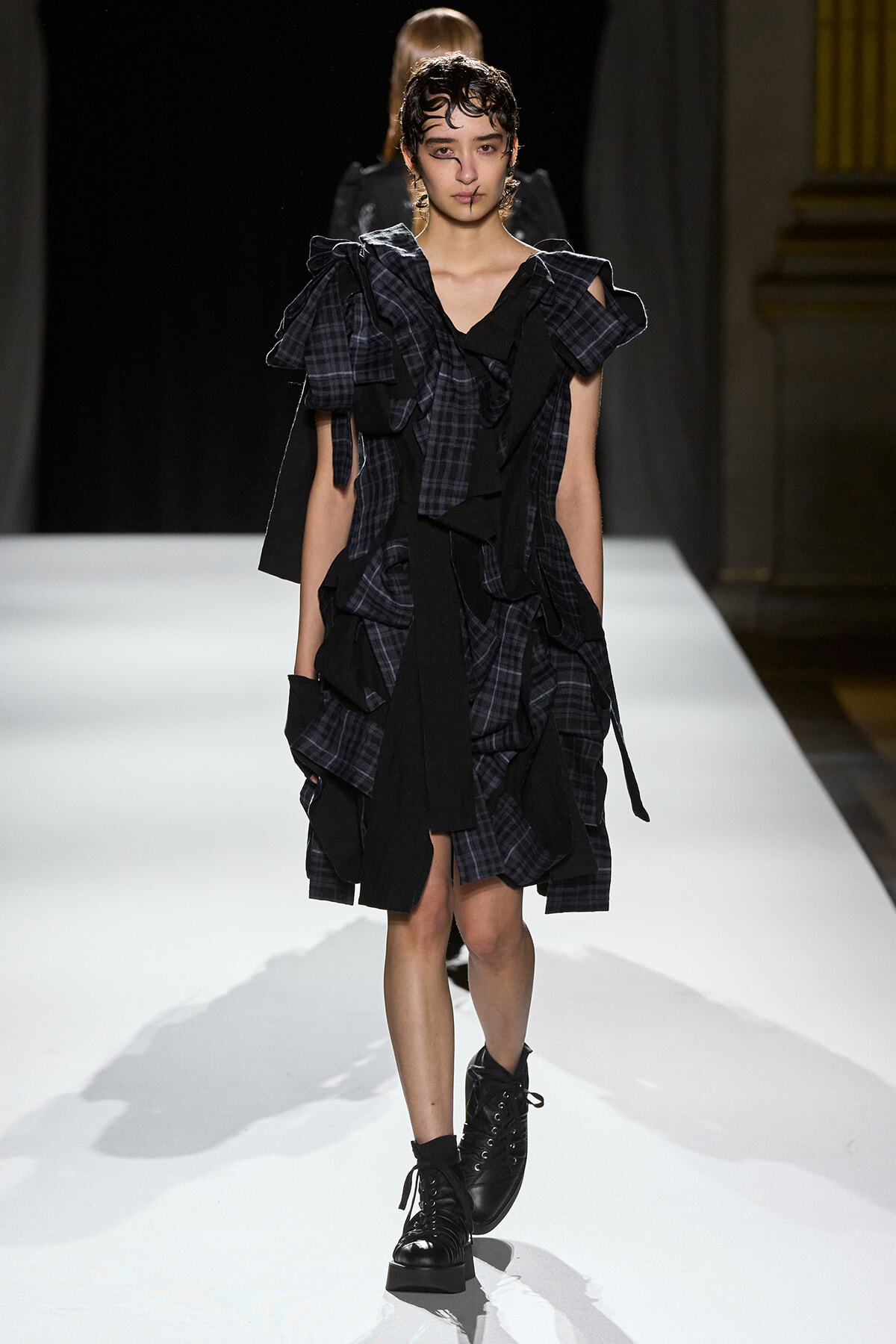 Model walks the runway in a dark plaid, ruffled dress with oversized shoulders and black combat boots.