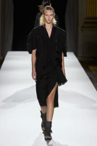 Model walking a white runway in a black, layered dress with puff sleeves and fringed details.