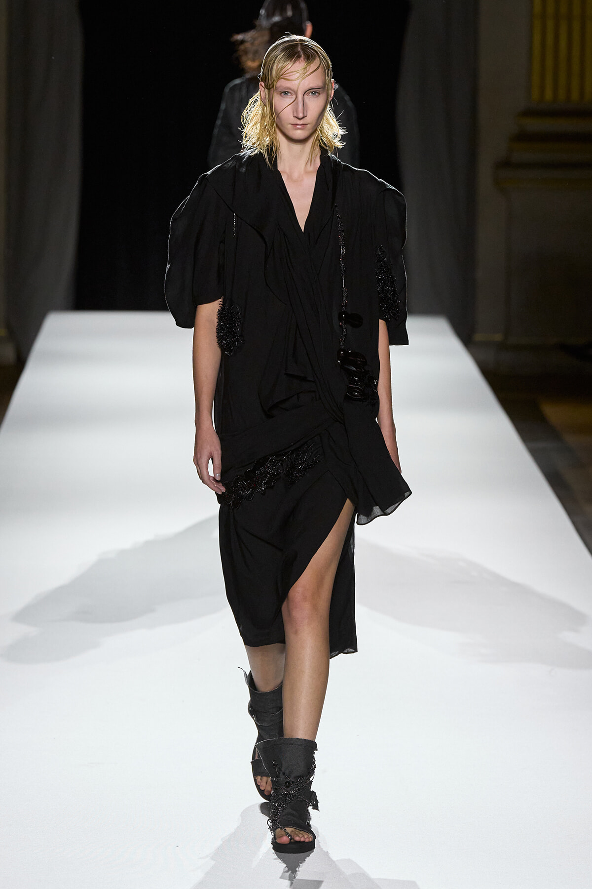 Model walking a white runway in a black, layered dress with puff sleeves and fringed details.