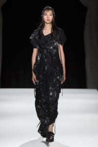 Model walks a black, textured, ruffled dress with tie details on a white runway.