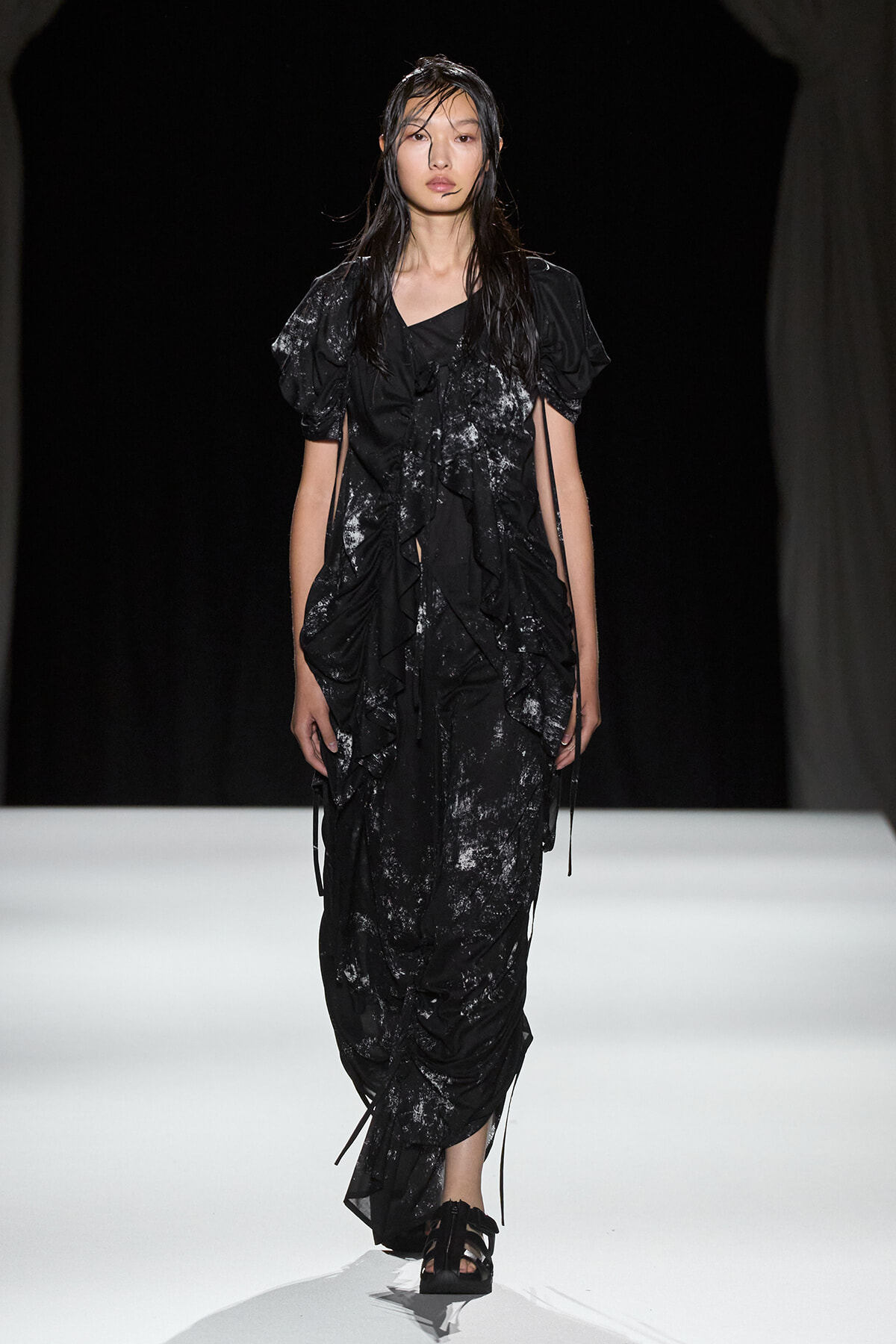 Model walks a black, textured, ruffled dress with tie details on a white runway.