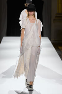 Model walks a white, sheer, layered gown with draped panels on a fashion runway, wearing a black cap and gloves.