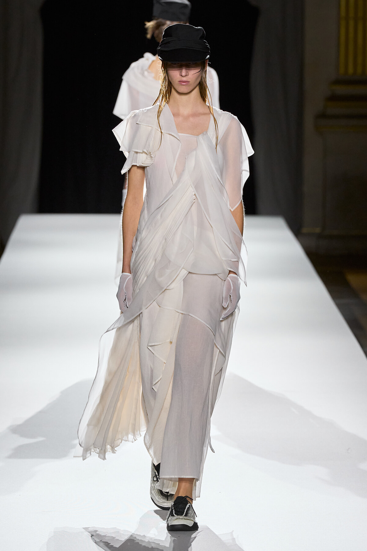 Model walks a white, sheer, layered gown with draped panels on a fashion runway, wearing a black cap and gloves.