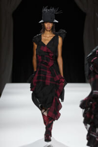 Runway model in a black and burgundy ruffled wrap dress, paired with a tall black hat featuring feathered accents.