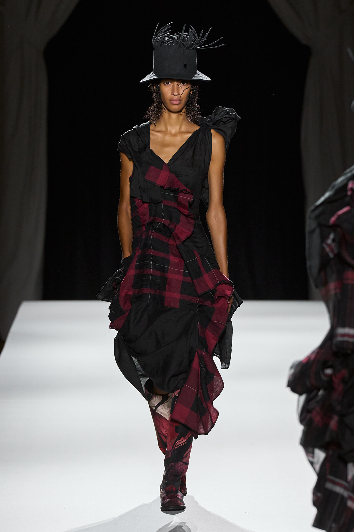 Runway model in a black and burgundy ruffled wrap dress, paired with a tall black hat featuring feathered accents.